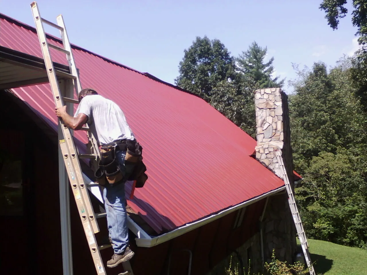 Roofer on a ladder beside a red metal roof and stone chimney — Soffit & Fascia Repair by Roofing Brothers in Purdys
